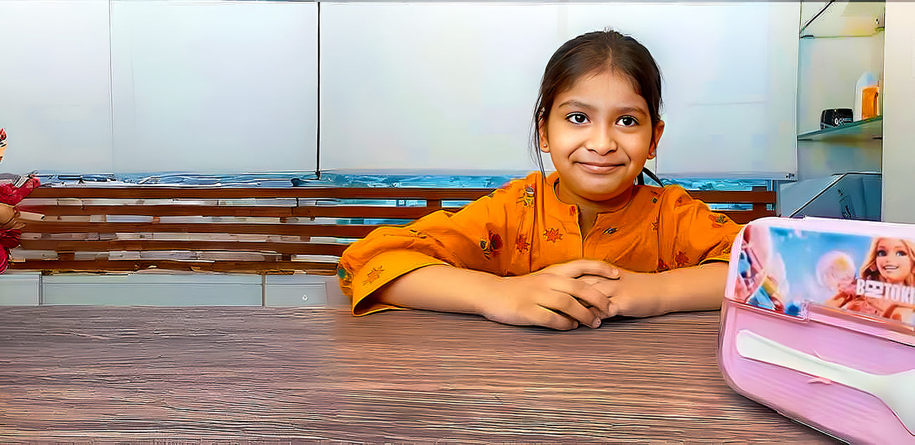 Load video: Musfira Bilal, the CEO of Bentokids, a young girl with a bright smile, sitting at a wooden table in an orange shirt. A pink Bentokids lunchbox with a doll graphic is visible on the right. Is this video where Musfira Bilal is describing the features of the Bentokids product