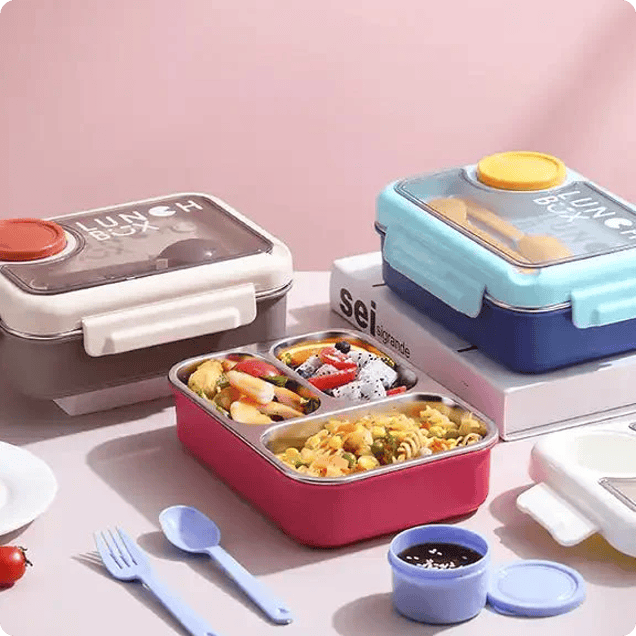 Close-up of a pink bento-style lunchbox with three compartments filled with food, including pasta, shrimp, and fruit salad. A light blue spoon and fork, and a small sauce container are in the foreground.